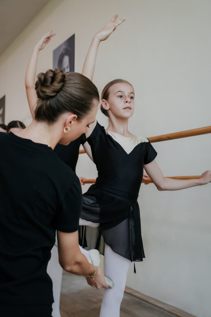 Ballet class with teacher assisting young girl in stretching at barre, wearing black leotard and white tights.