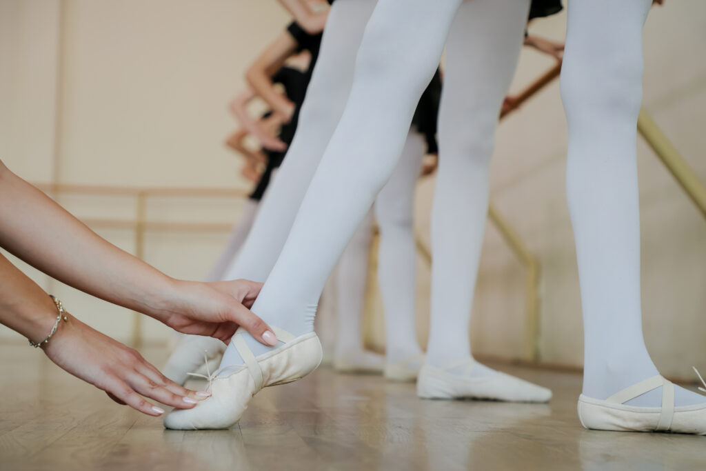 Close-up of ballet teacher correcting foot position of young student in white tights and ballet slippers during class.